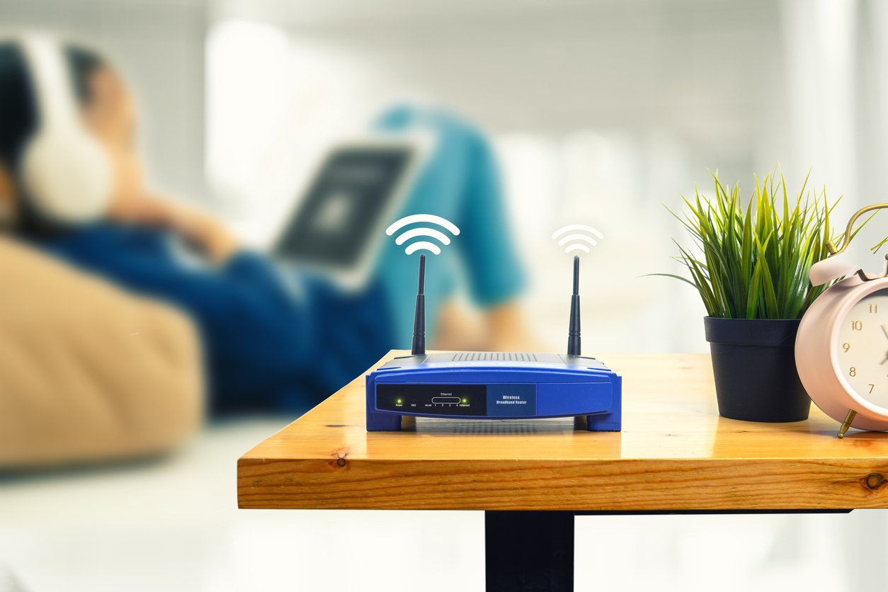 Router with WiFi signal above antennae on side table as someone watches an iPad in the background.