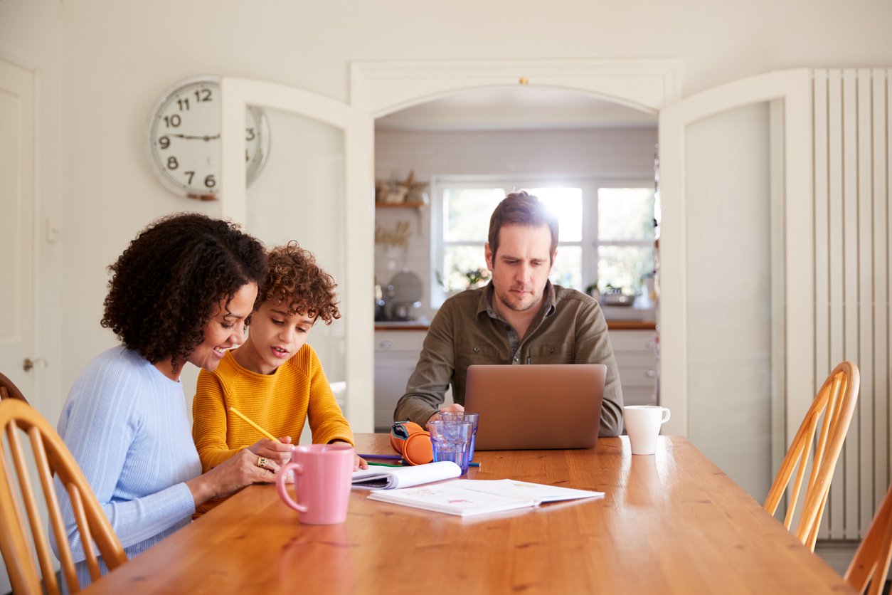 A father works from the kitchen table while a mother helps their son with schoolwork.