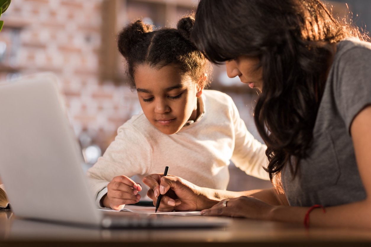 A mother helps her young daughter with schoolwork at home with a laptop in the foreground. 
