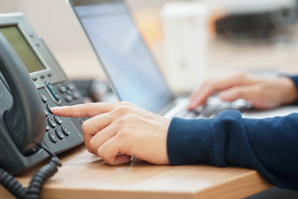 Person dialing the number pad on a desk phone while on a laptop.