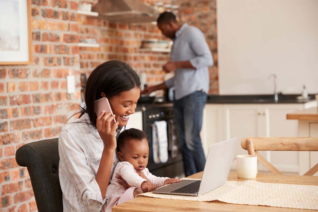 Woman on the phone with a baby in her lap, working remotely at her kitchen table.