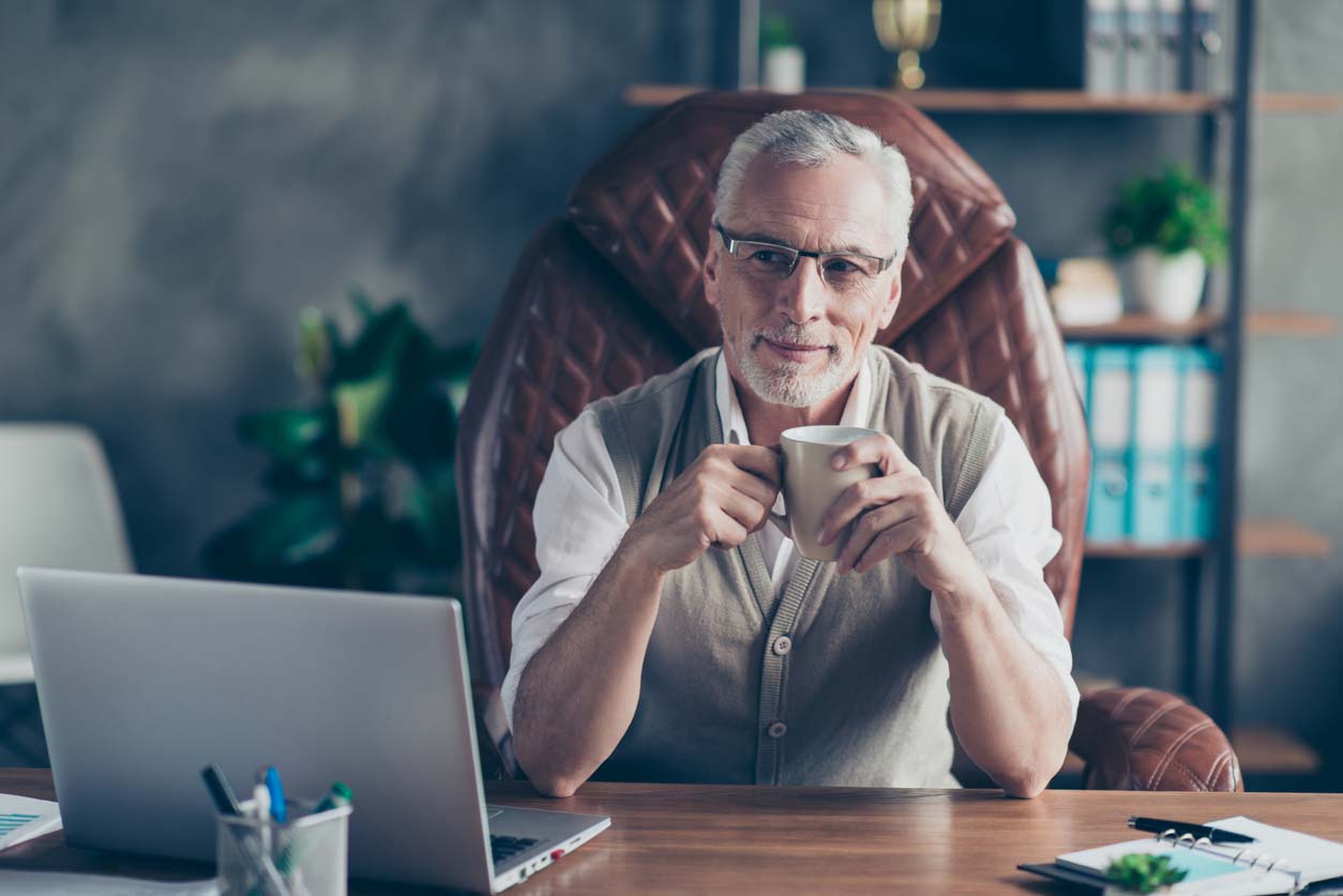 Retirement-age man sits at his work desk sipping coffee.